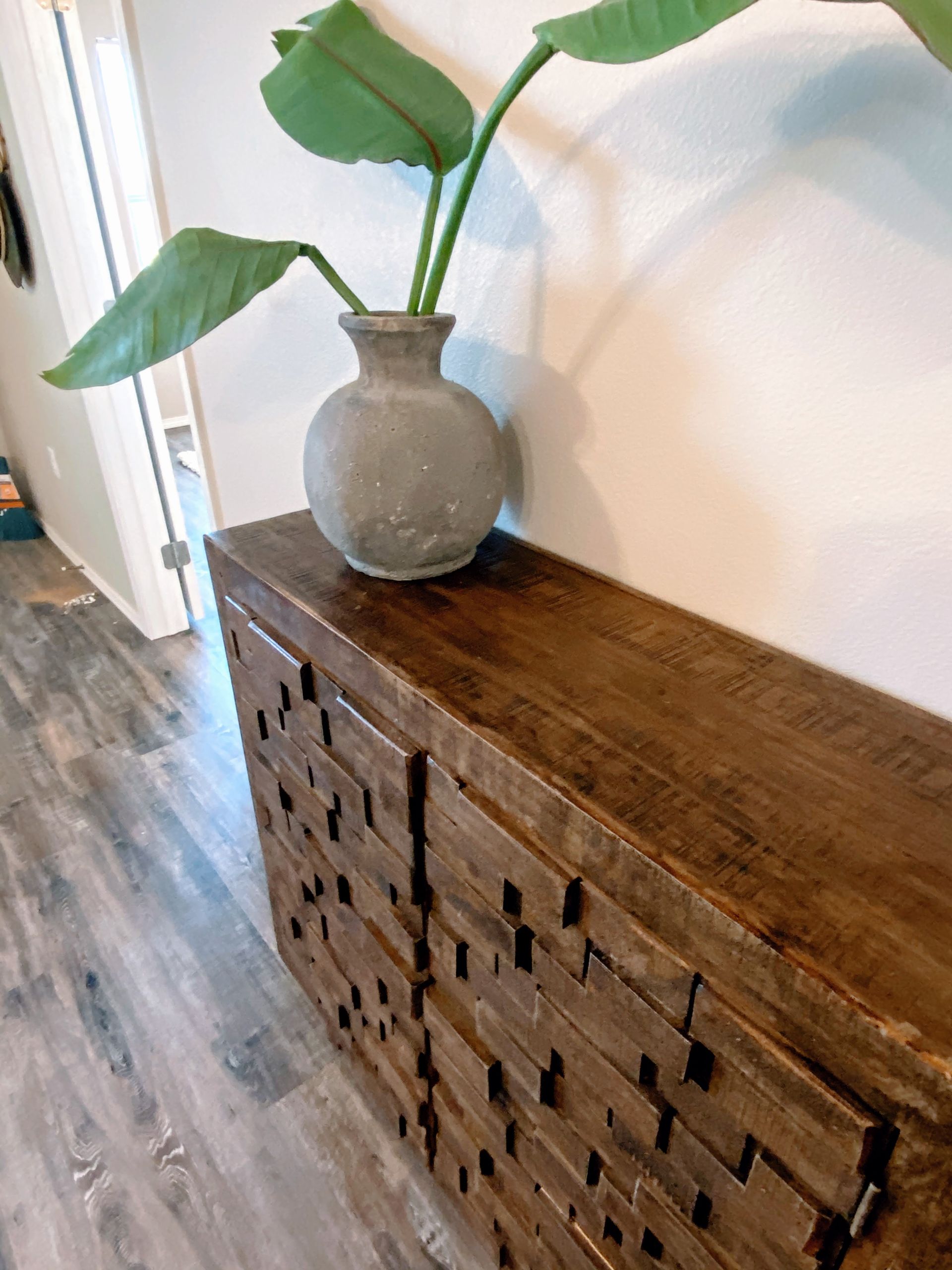 Wooden console table with textured front, topped with a gray vase holding green plant stems, against a white wall.