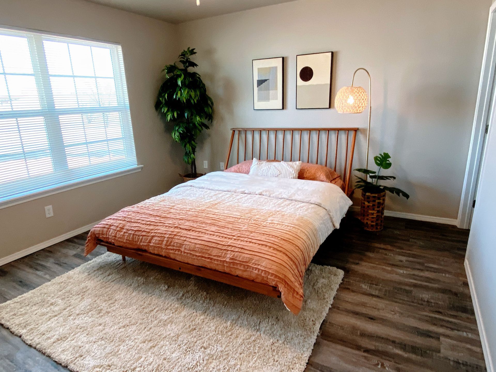Bedroom with wooden bed, rug, plants, and art. Neutral walls and flooring, natural light.