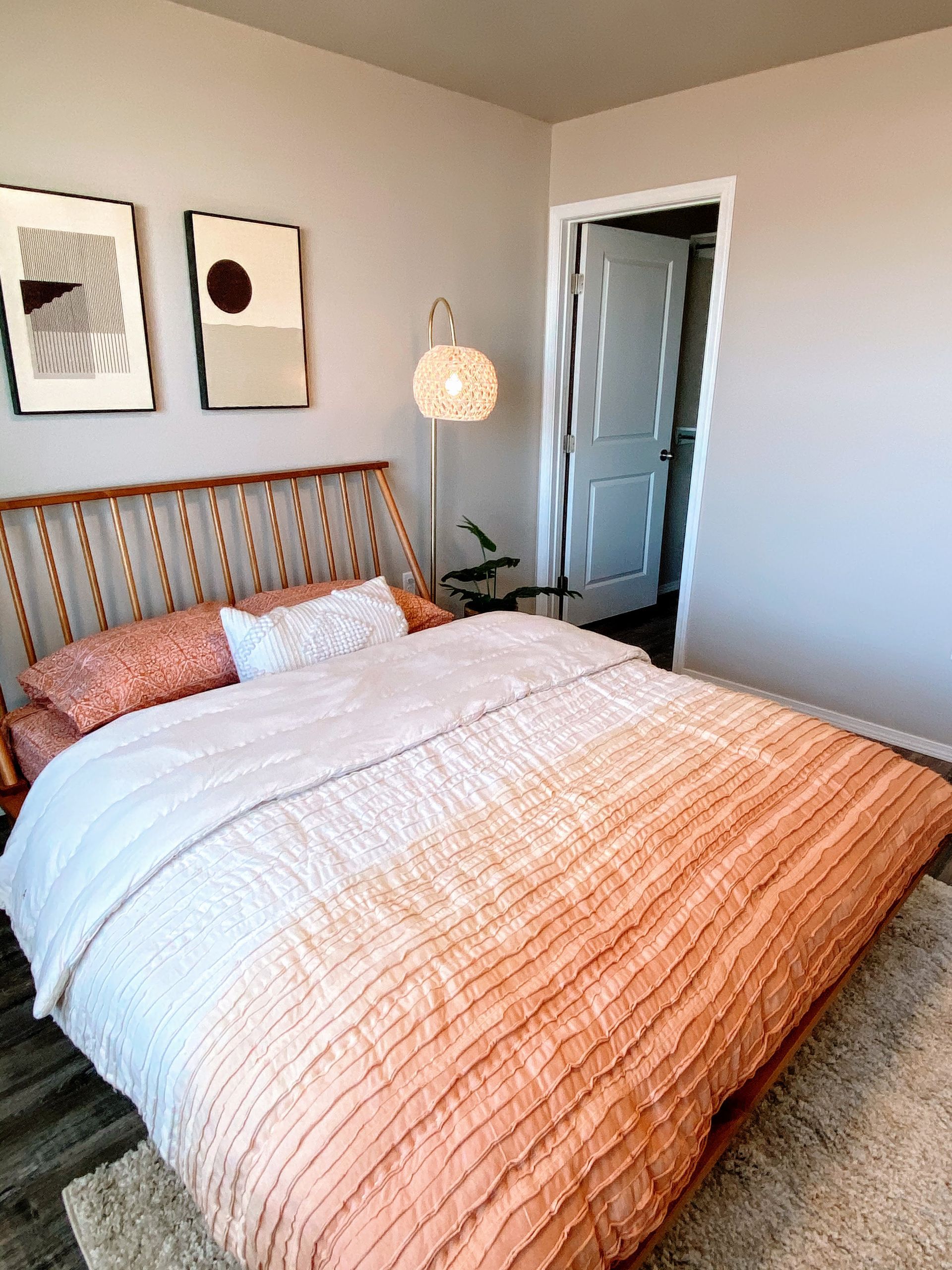Bedroom with orange ombre bedding, wooden headboard, art prints, and a woven floor lamp.