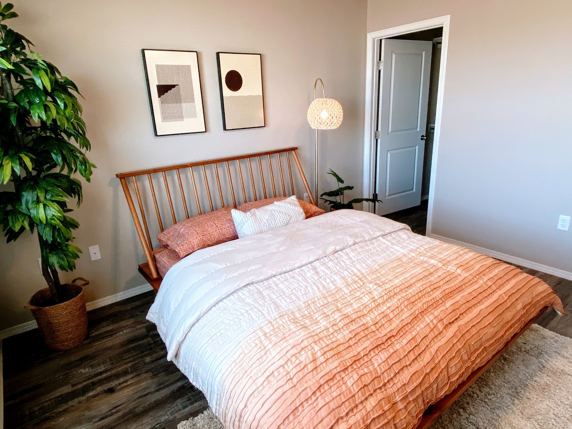 Bedroom with a wooden headboard, coral bedding, and a large potted plant. Two abstract art prints hang above the bed.