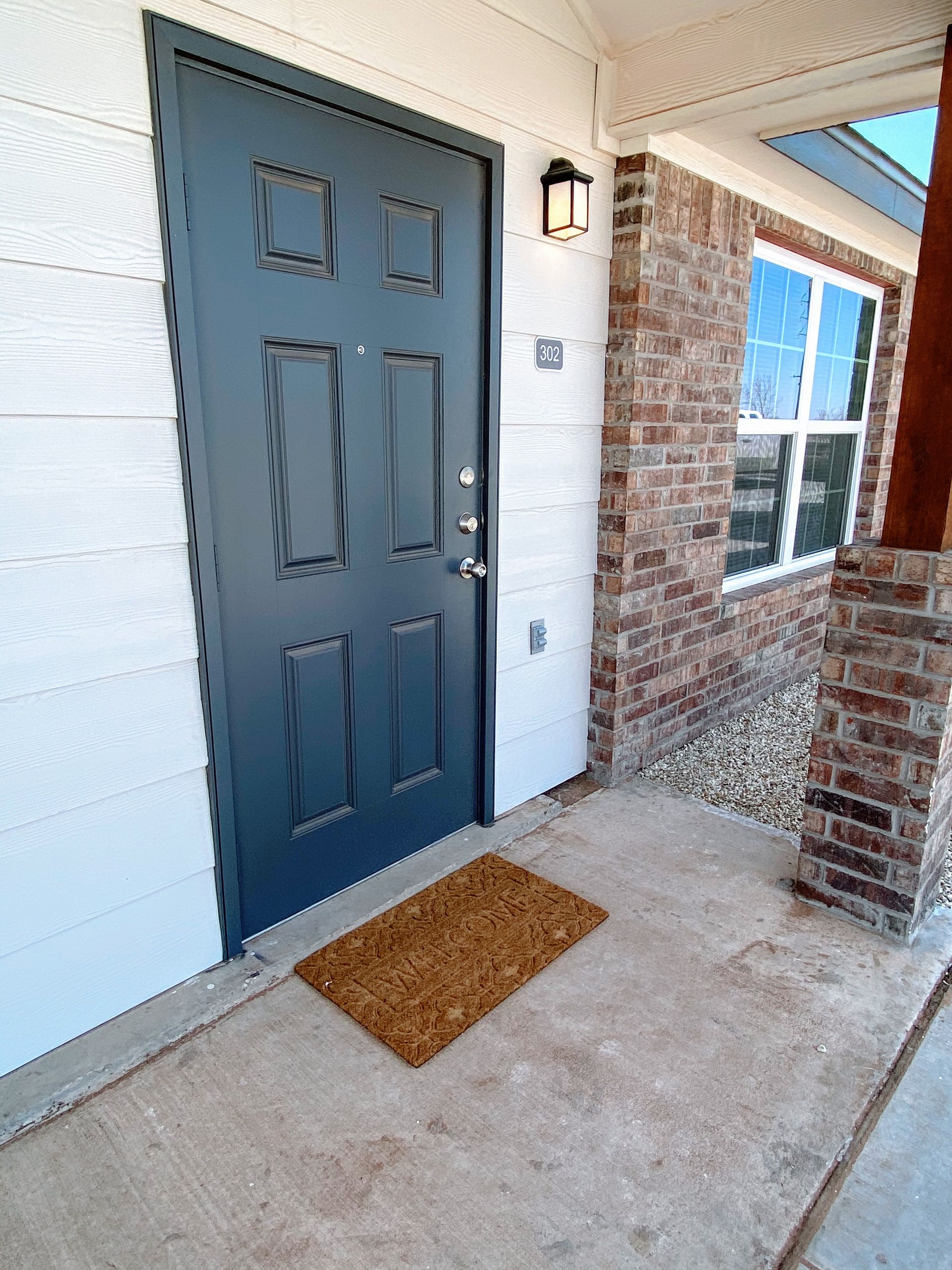 Blue front door with doormat, brick and white siding, and window.