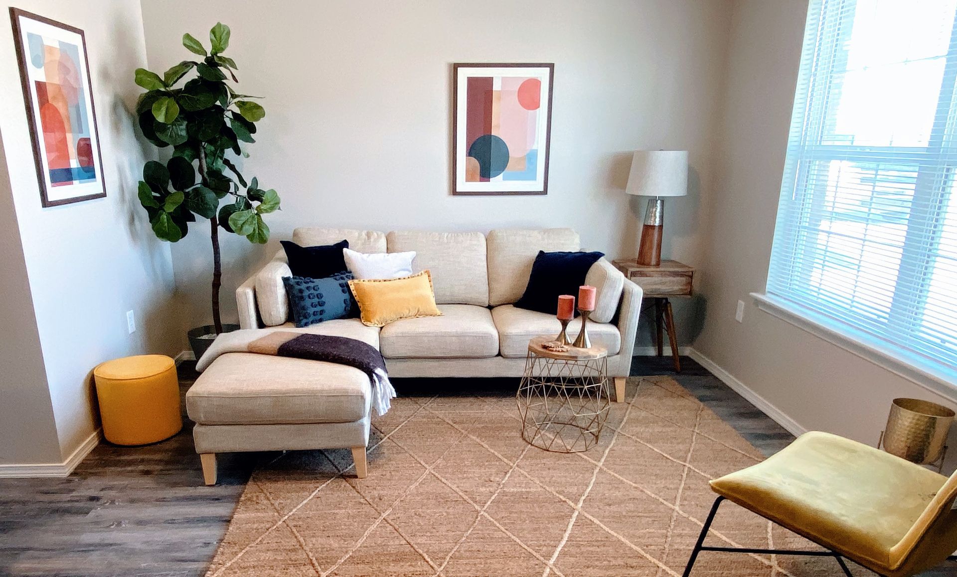Living room with beige sectional sofa, patterned rug, artwork, and tall plant.