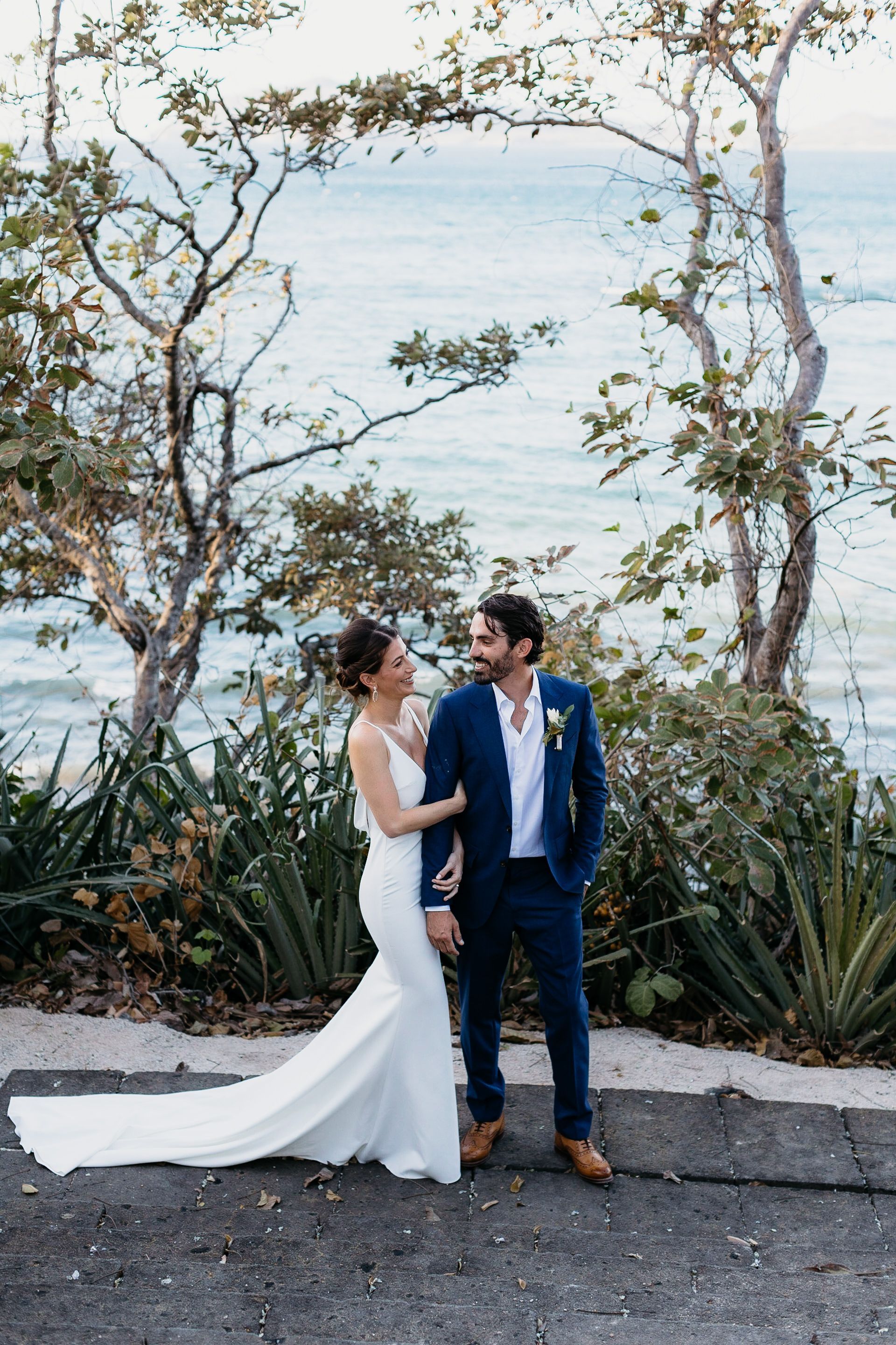 A bride and groom are standing next to each other in front of the ocean.