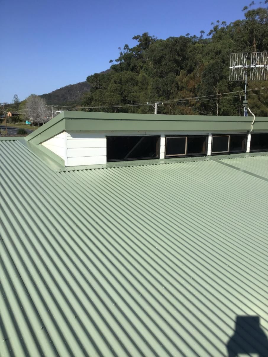 A Green Corrugated Metal Roof With A White Building In The Background — Glenroofing Pty Ltd In Wingham, NSW