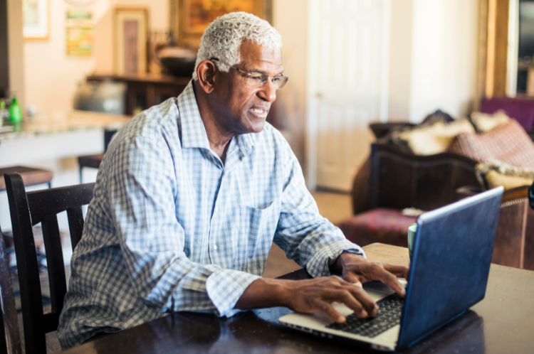 An older man is sitting at a table using a laptop computer.