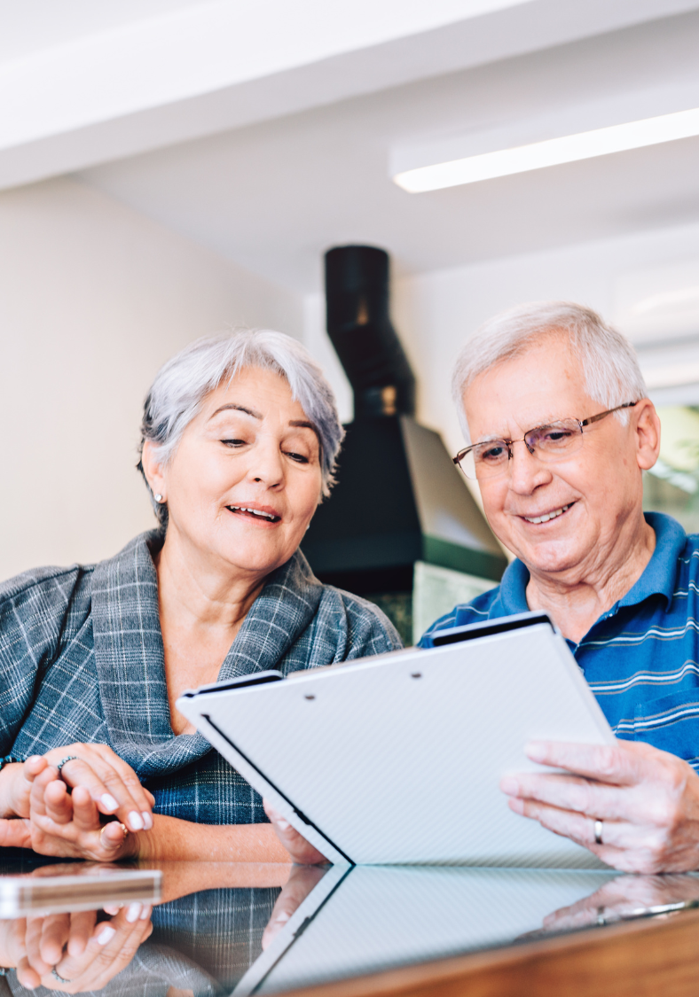 An elderly couple is sitting at a table looking at a tablet.