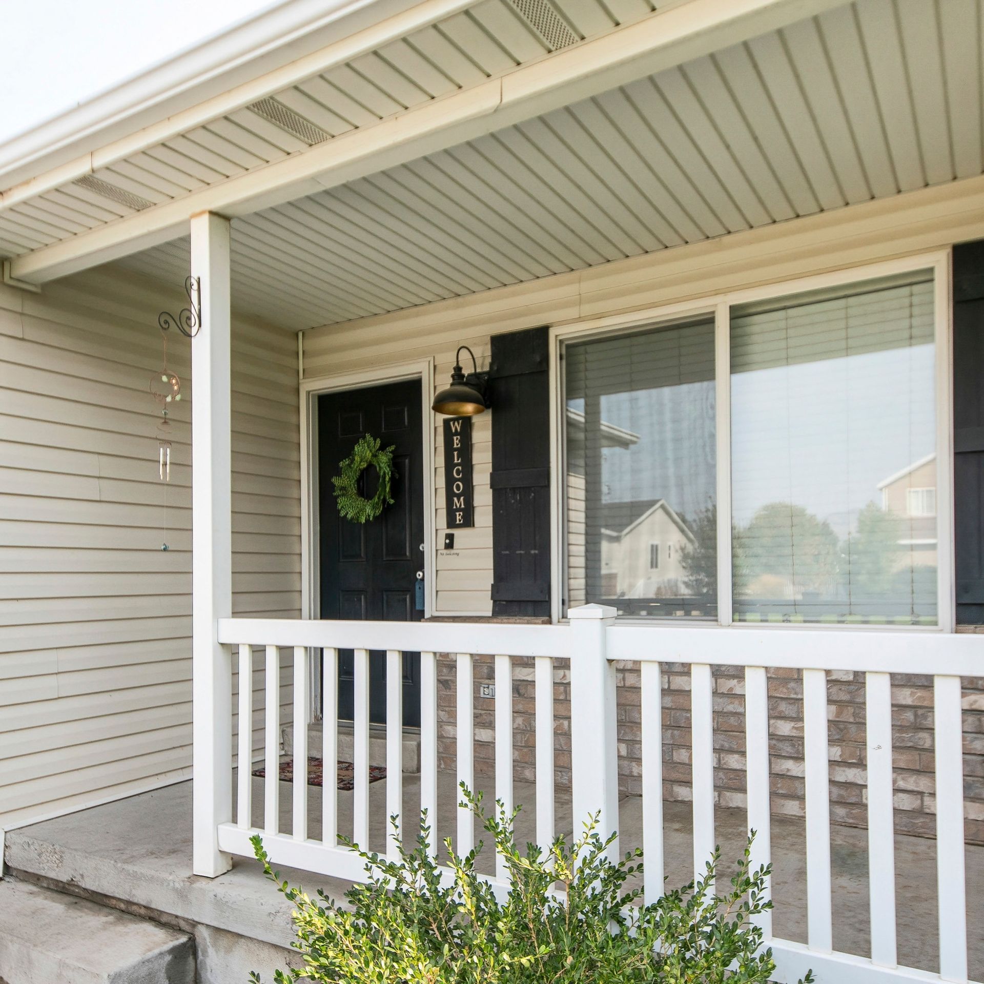 A beige house exterior with a covered porch, white railing, black front door with a wreath, and a large window.
