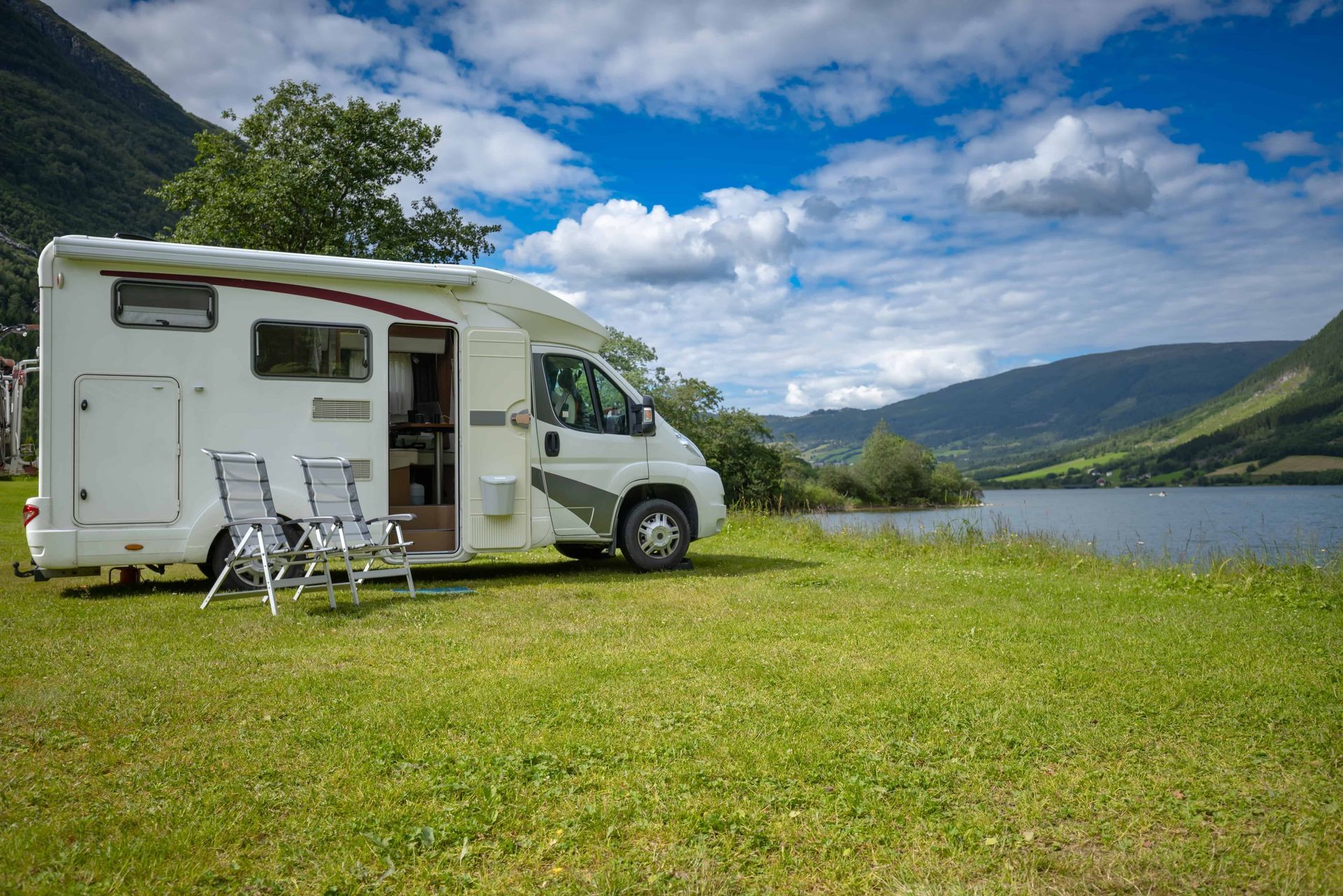 A white motorhome parked on a grassy riverbank near mountains under a blue, cloudy sky.