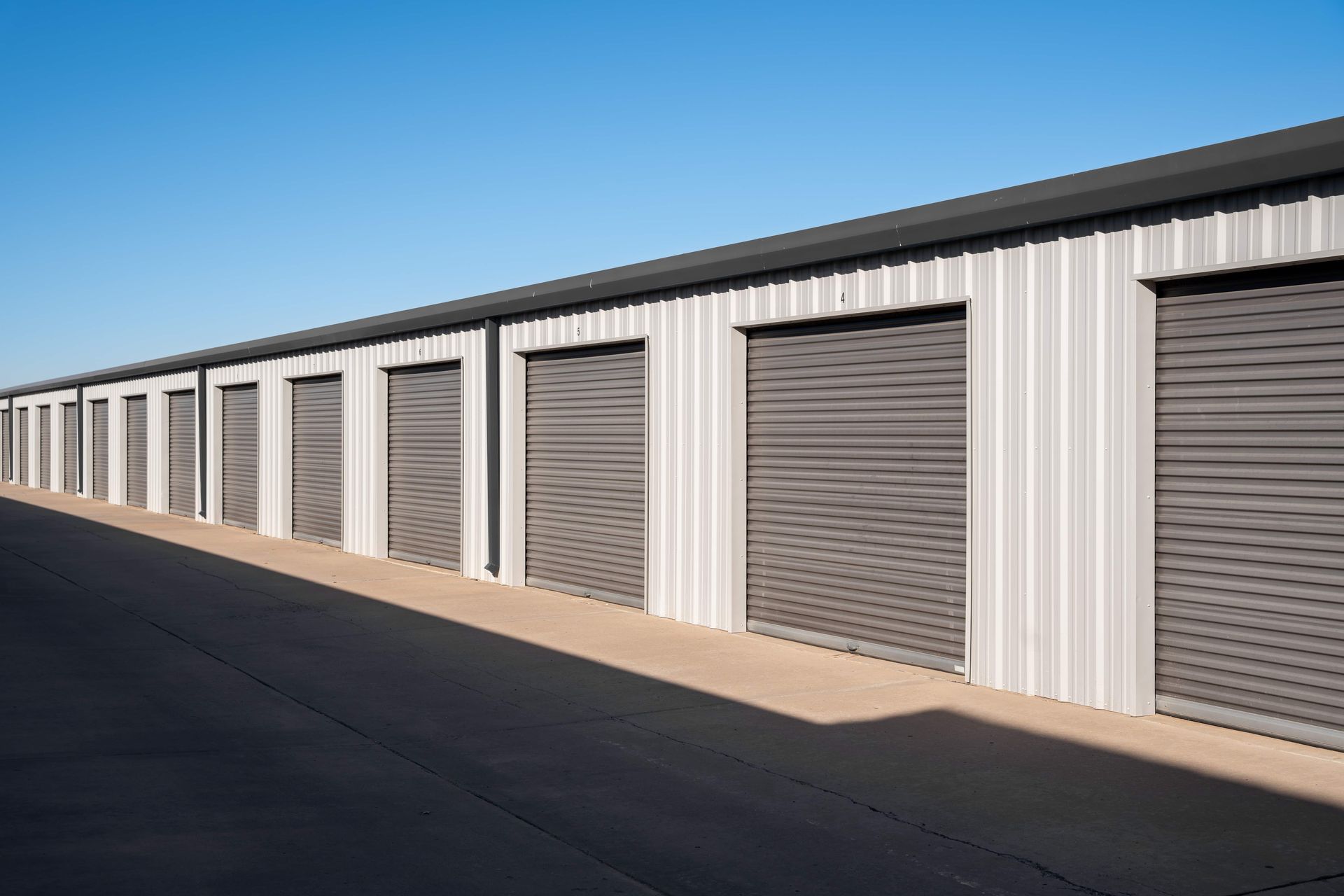 A row of light grey storage units with closed, dark grey roll-up doors against a clear blue sky.