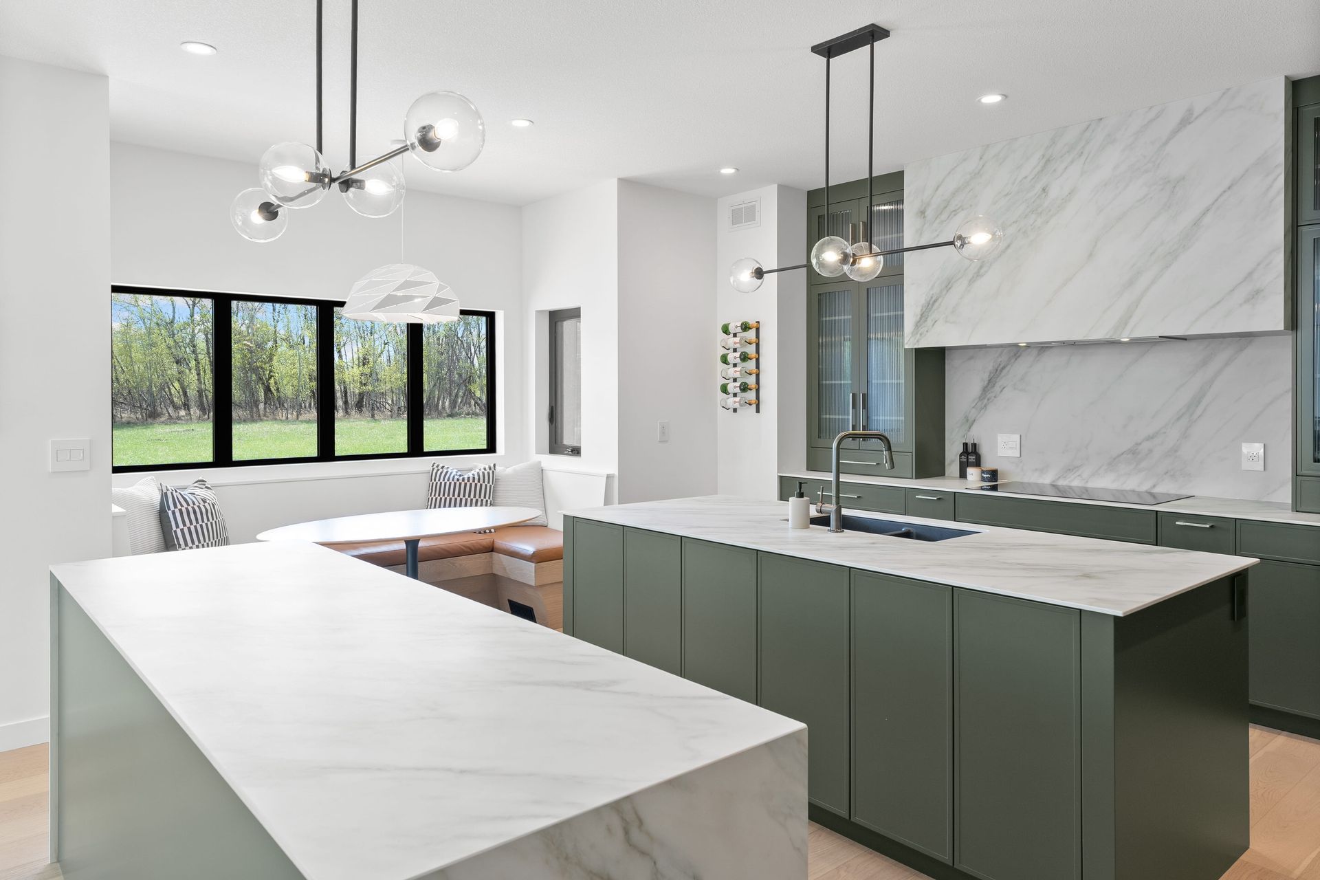 Modern kitchen with olive green cabinets, white marble islands, and a dining nook featuring a large window.