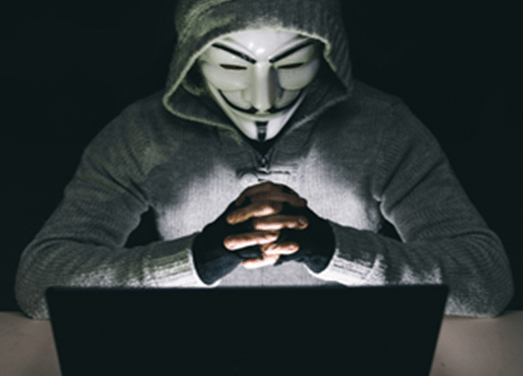 A man wearing a mask and a hoodie is looking at a laptop
