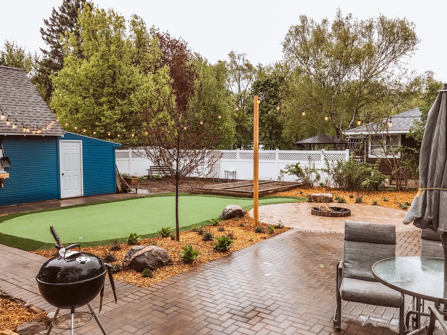 A backyard with a putting green , grill , table and chairs.