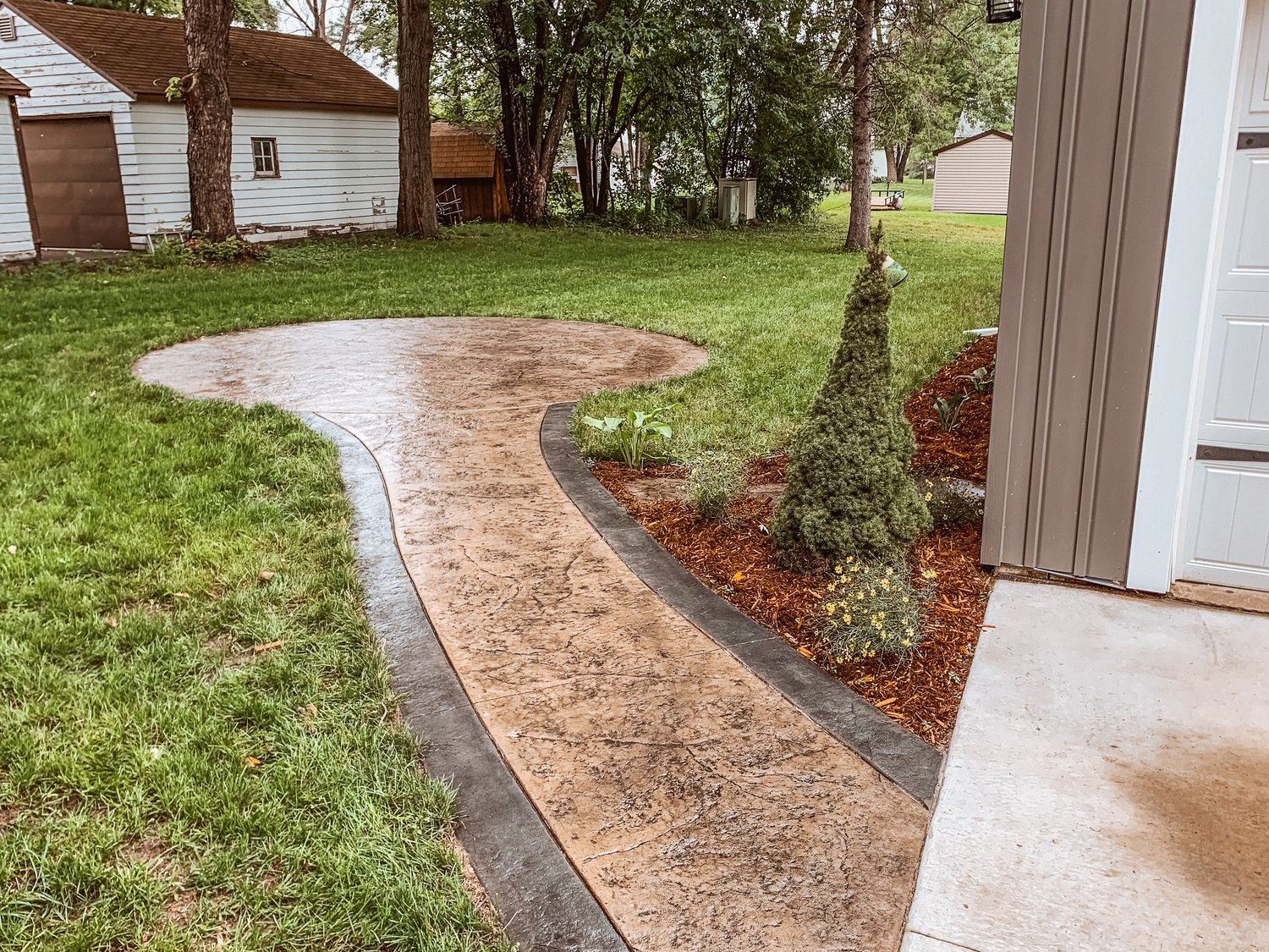 A concrete walkway leading to a house with a shed in the background.