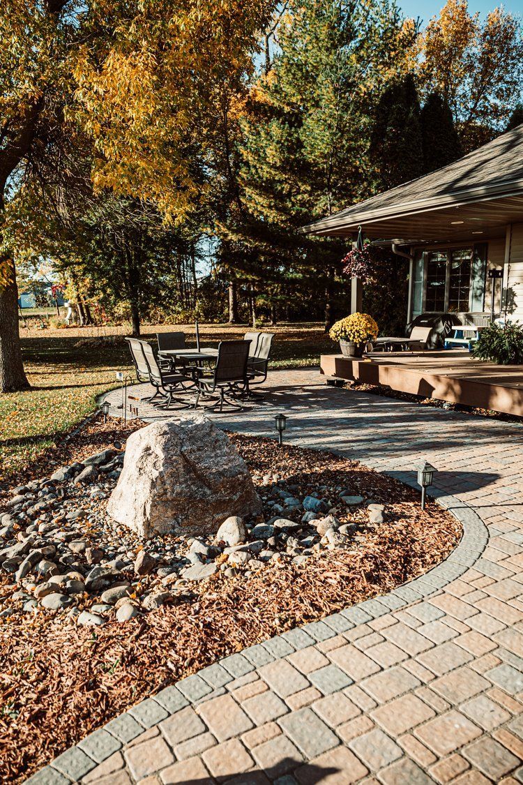 A brick walkway leading to a house with a patio and trees in the background.
