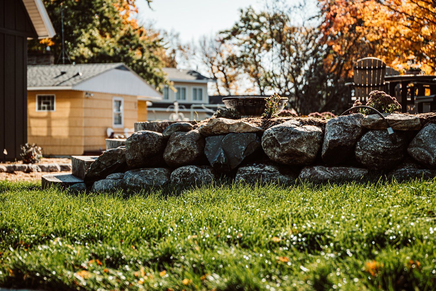 A large rock wall in a lush green yard with a house in the background.