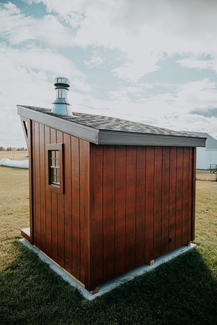 A small wooden shed with a chimney on the roof is sitting in the middle of a grassy field.