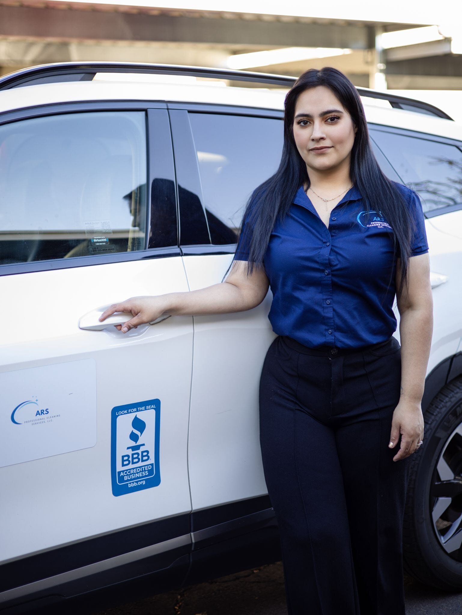 A woman in a blue shirt is standing next to a white car.