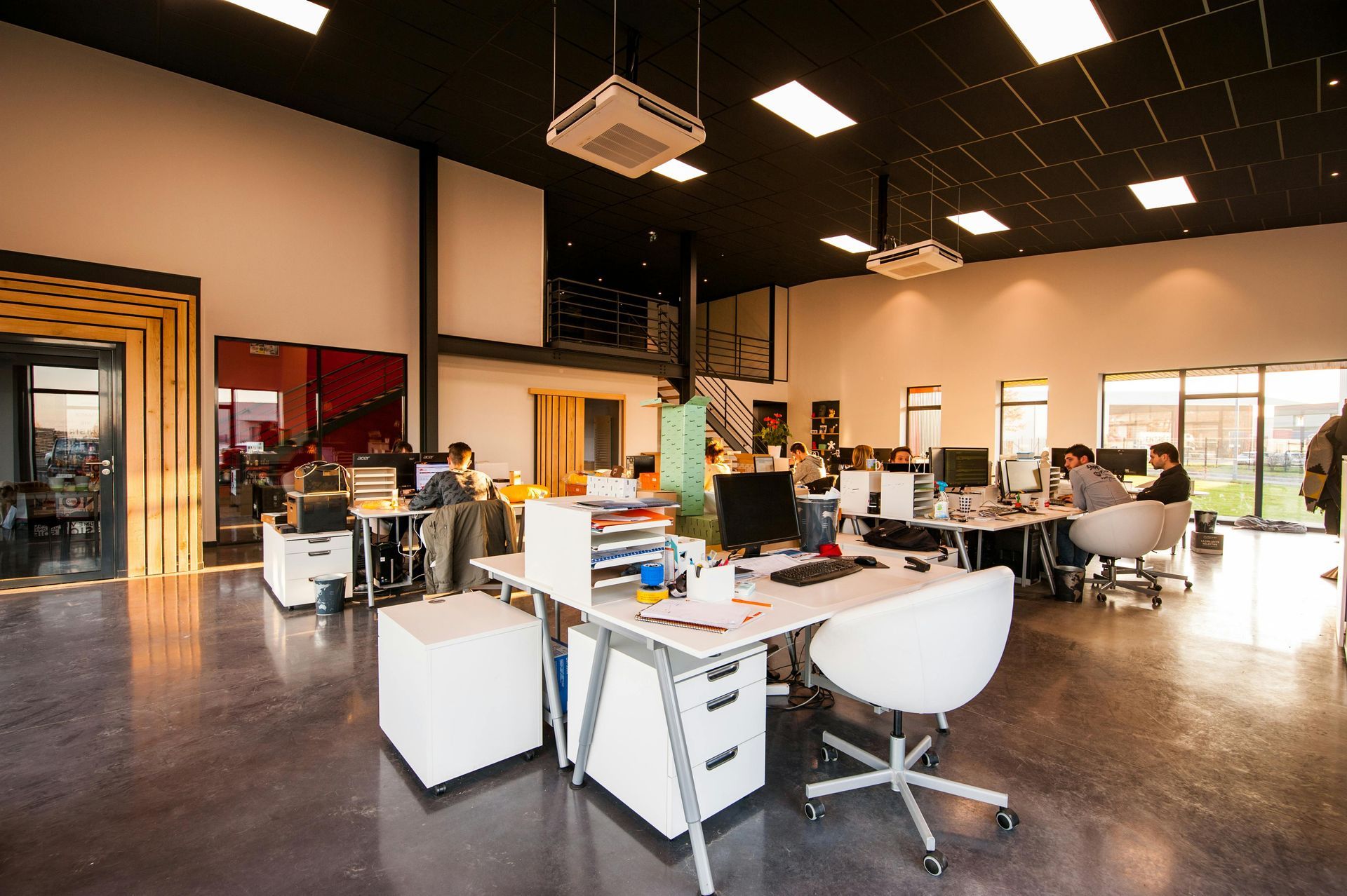 Open-plan office with desks, computers, chairs, and a few people working under high ceilings.