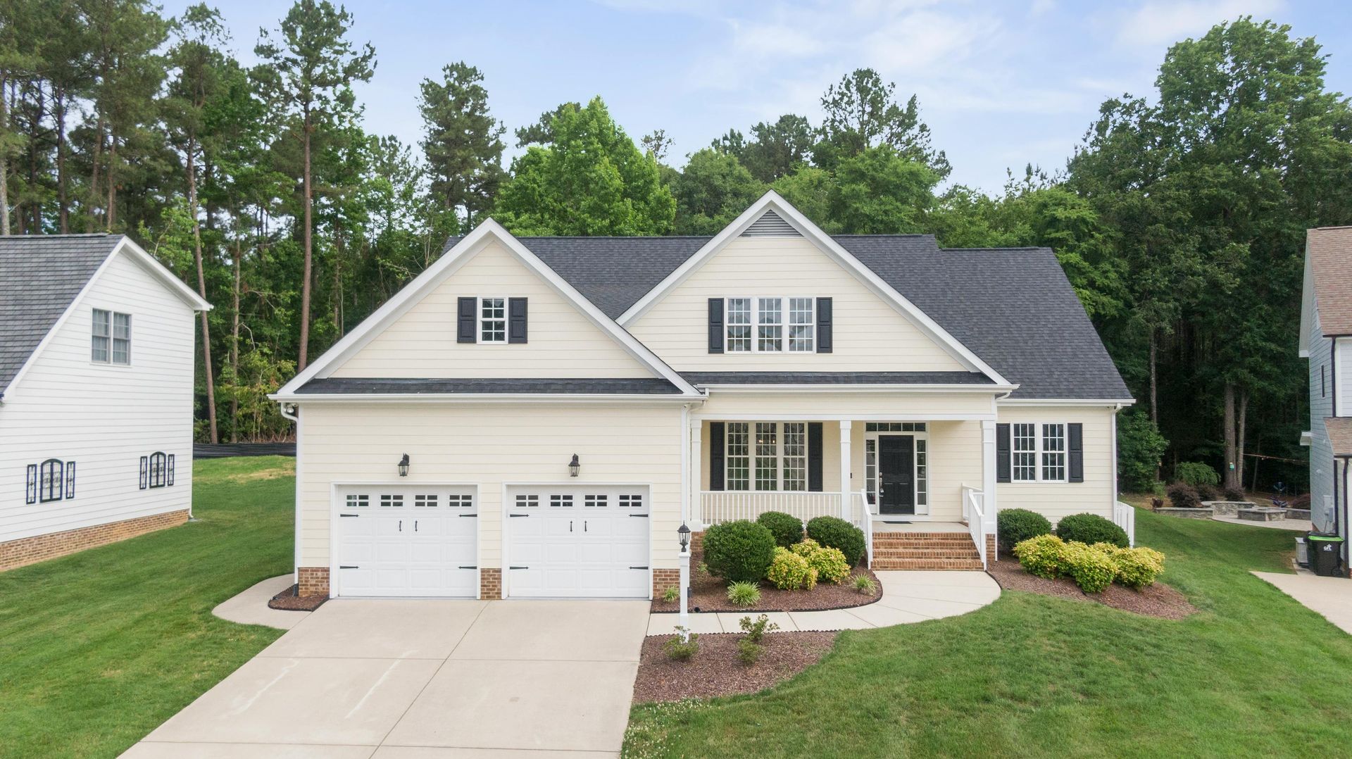 Suburban house with cream siding, two-car garage, front porch, and landscaped lawn under trees