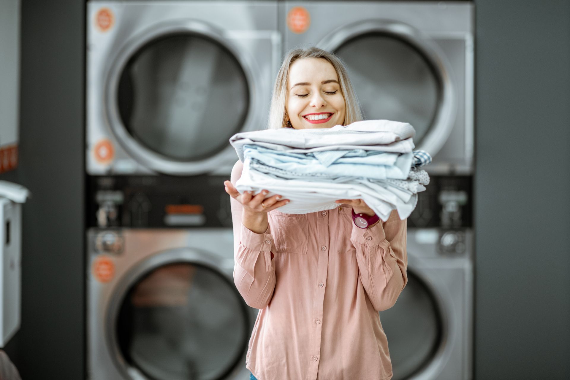 A woman holding clean clothes in the self-service laundry with dryer machines in the background.
