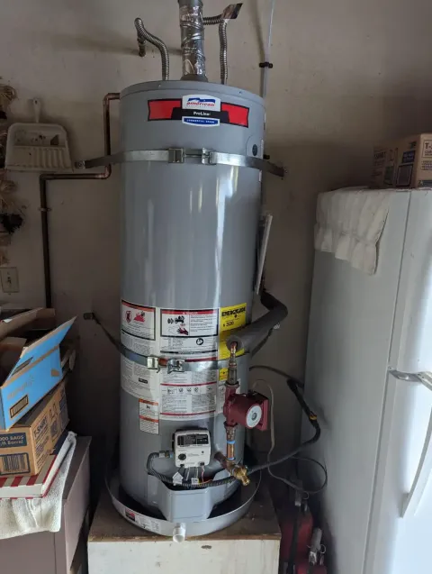 Gray water heater in a utility closet with pipes, labels, and a white refrigerator beside it