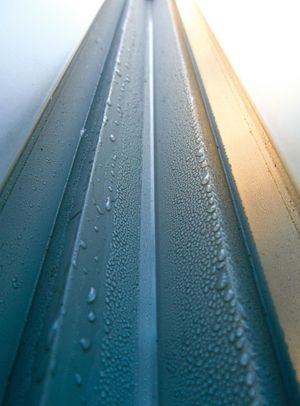 Close-up of a blue metal surface with vertical grooves, covered in water droplets. Sunlight highlights one edge.