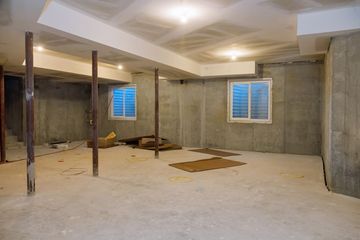 Unfinished basement with concrete walls, support beams, two small windows, and scattered materials.