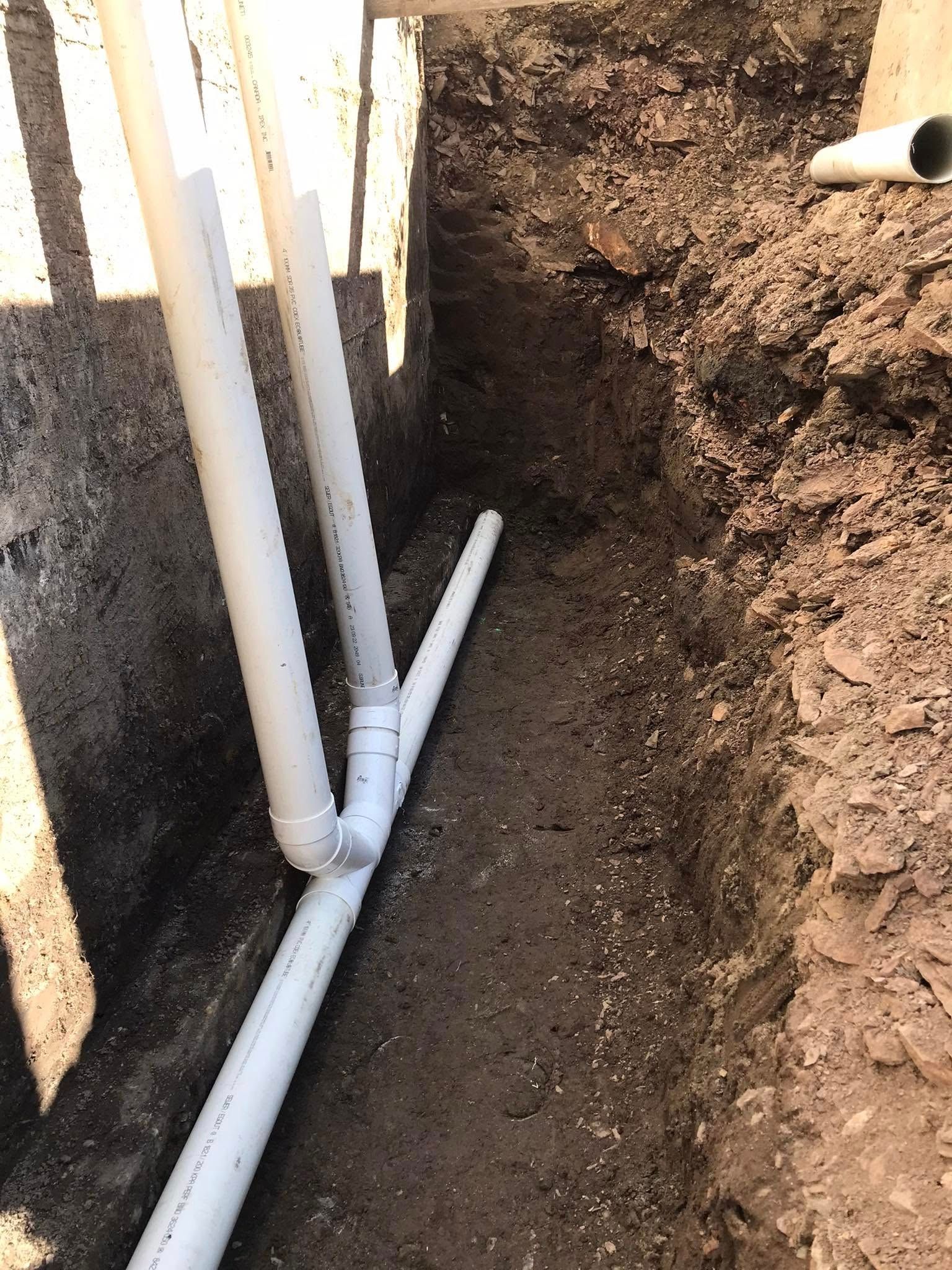 Tuyaux en PVC blanc dans une tranchée, raccordés. La terre entoure les tuyaux, et le soleil brille au-dessus.
