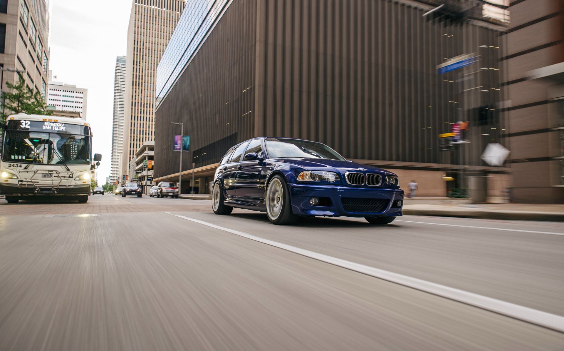 A blue car is driving down a city street next to a bus. | Import Specialty Service