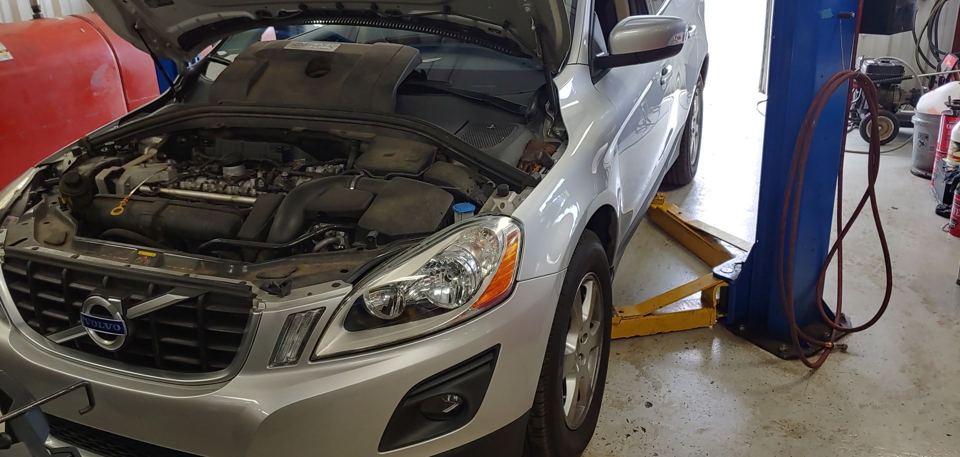 A silver volvo with the hood up is sitting on a lift in a garage. | Import Specialty Service