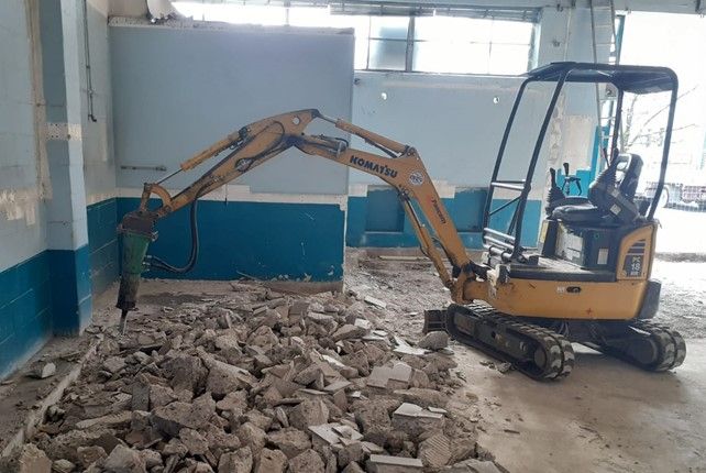Un escavatore giallo Komatsu rompe il cemento in un edificio con pareti blu.