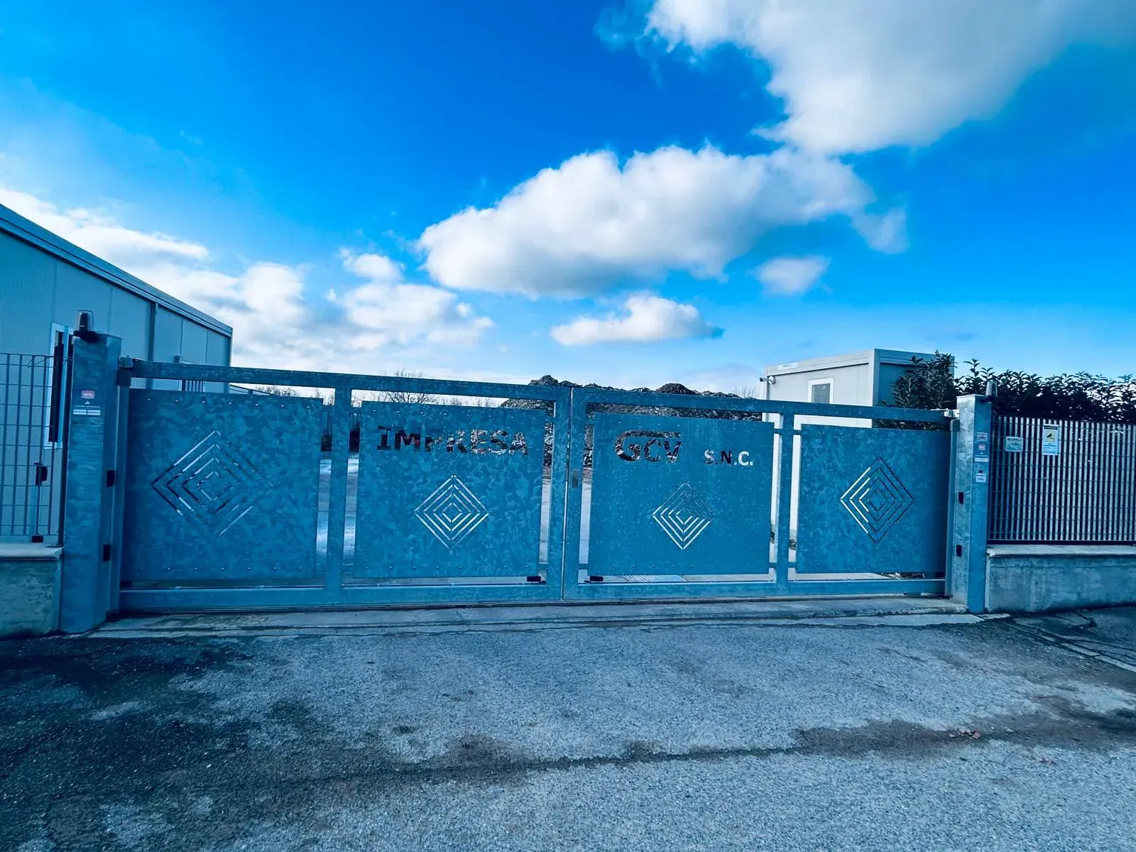 Grande cancello di metallo blu con disegni geometrici, sullo sfondo di un cielo nuvoloso.