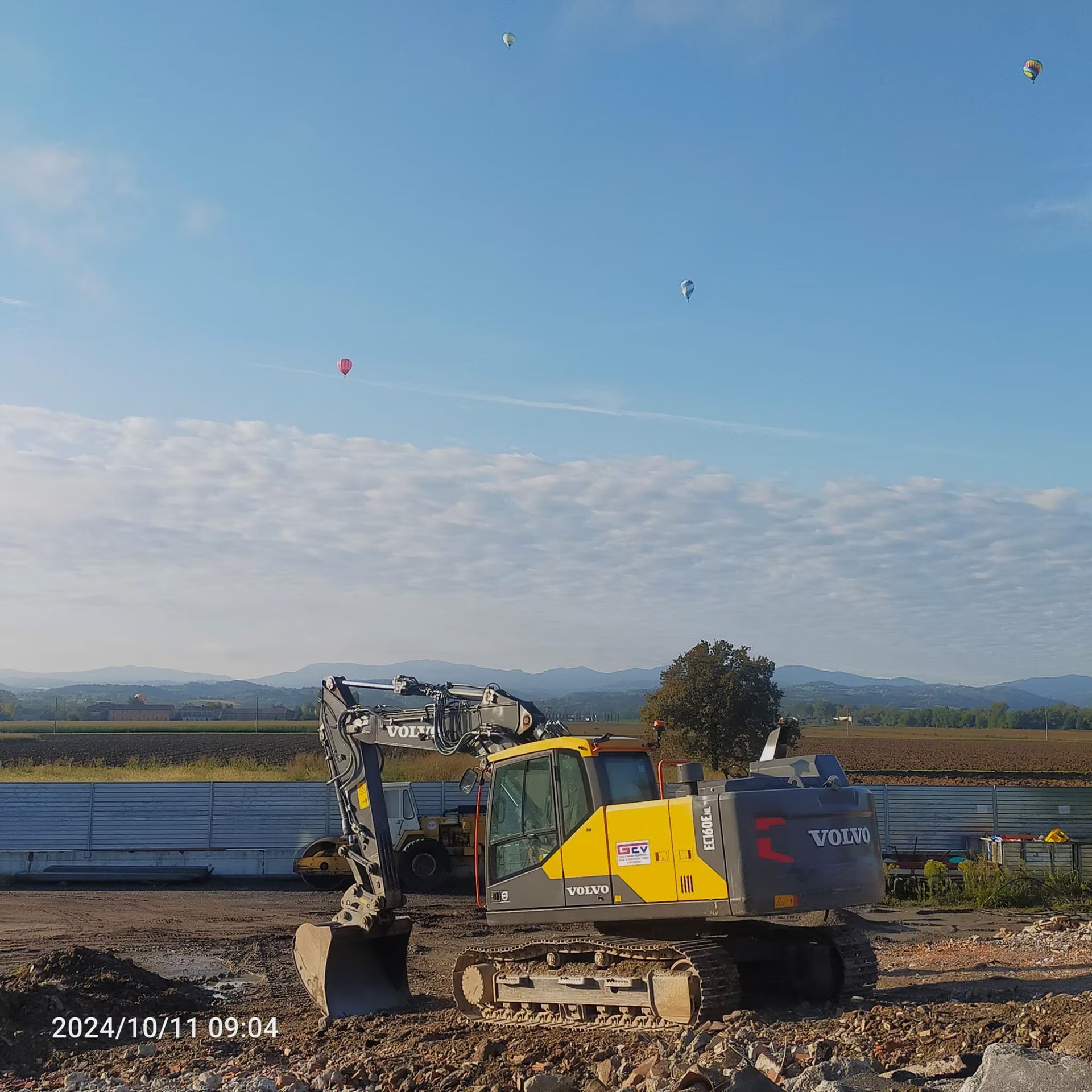 Escavatore in un cantiere con cielo azzurro e campi sullo sfondo.
