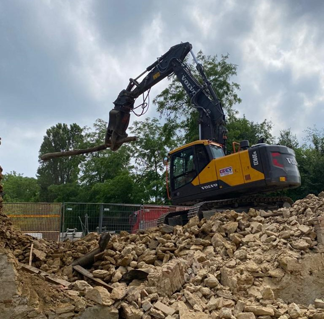 Un escavatore giallo demolisce una struttura in pietra all'aperto, sotto un cielo nuvoloso.
