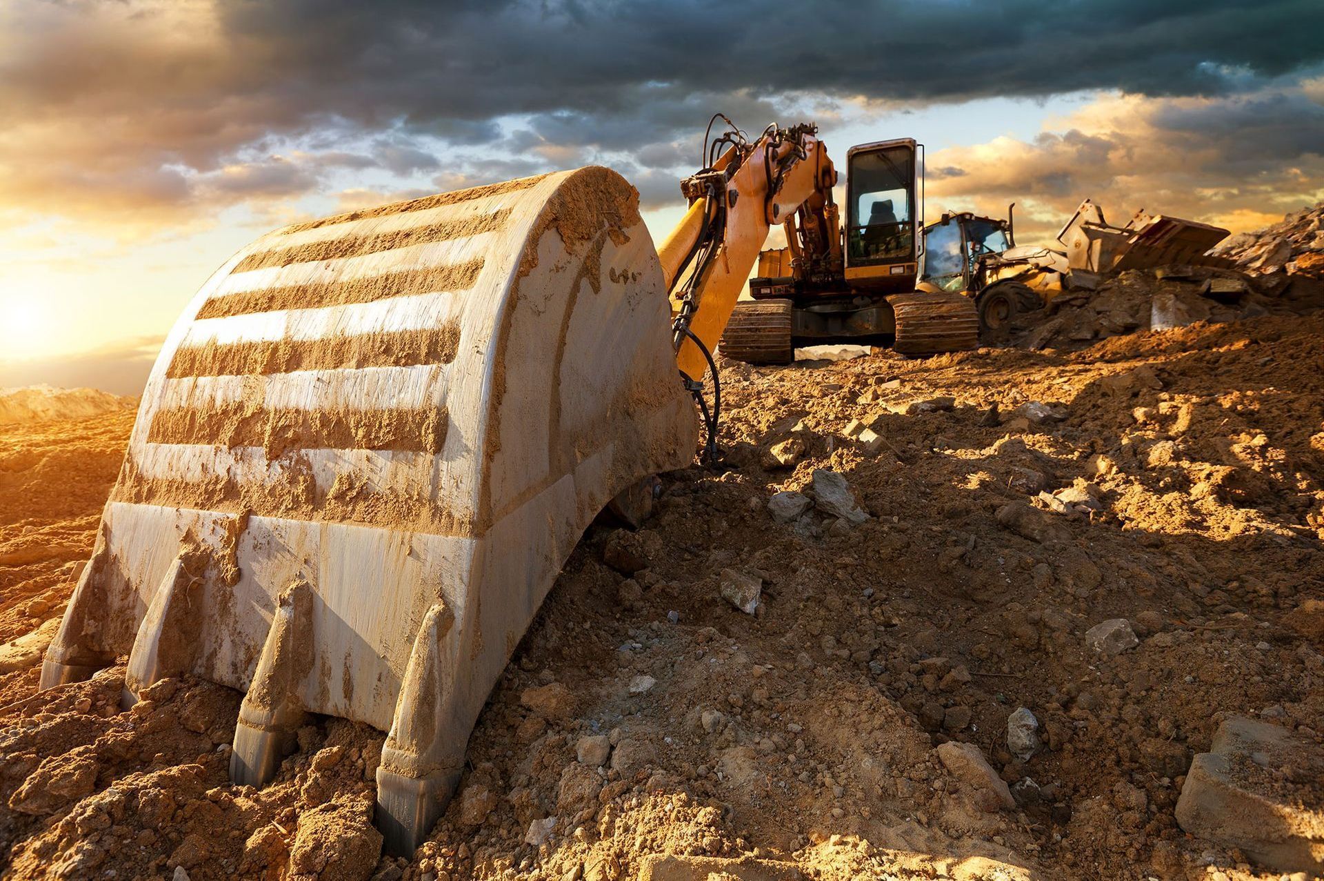 La benna di un escavatore raccoglie terra in un cantiere; cielo coperto e luce solare.