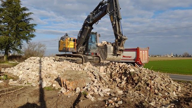 Un escavatore carica macerie su un autocarro con cassone ribaltabile, all'aperto in una giornata di sole.