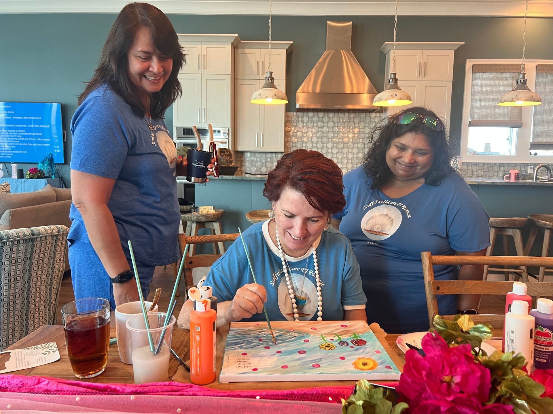 Three women are sitting at a table painting on a canvas.