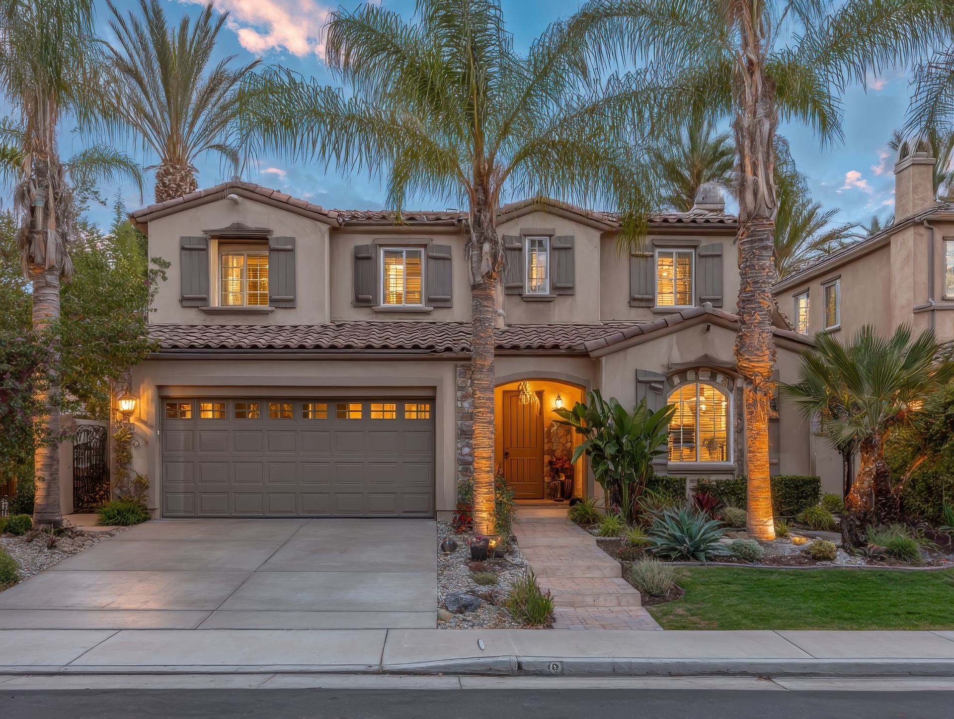 Beige two-story house with Spanish tile roof, arched windows, and palm trees. Front entrance with pathway lit by spotlights.
