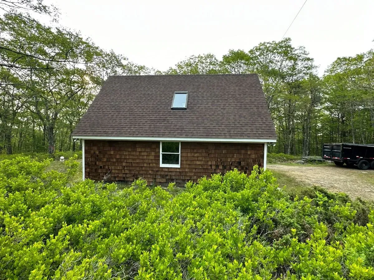 A small house with a skylight on the roof is surrounded by trees and bushes.