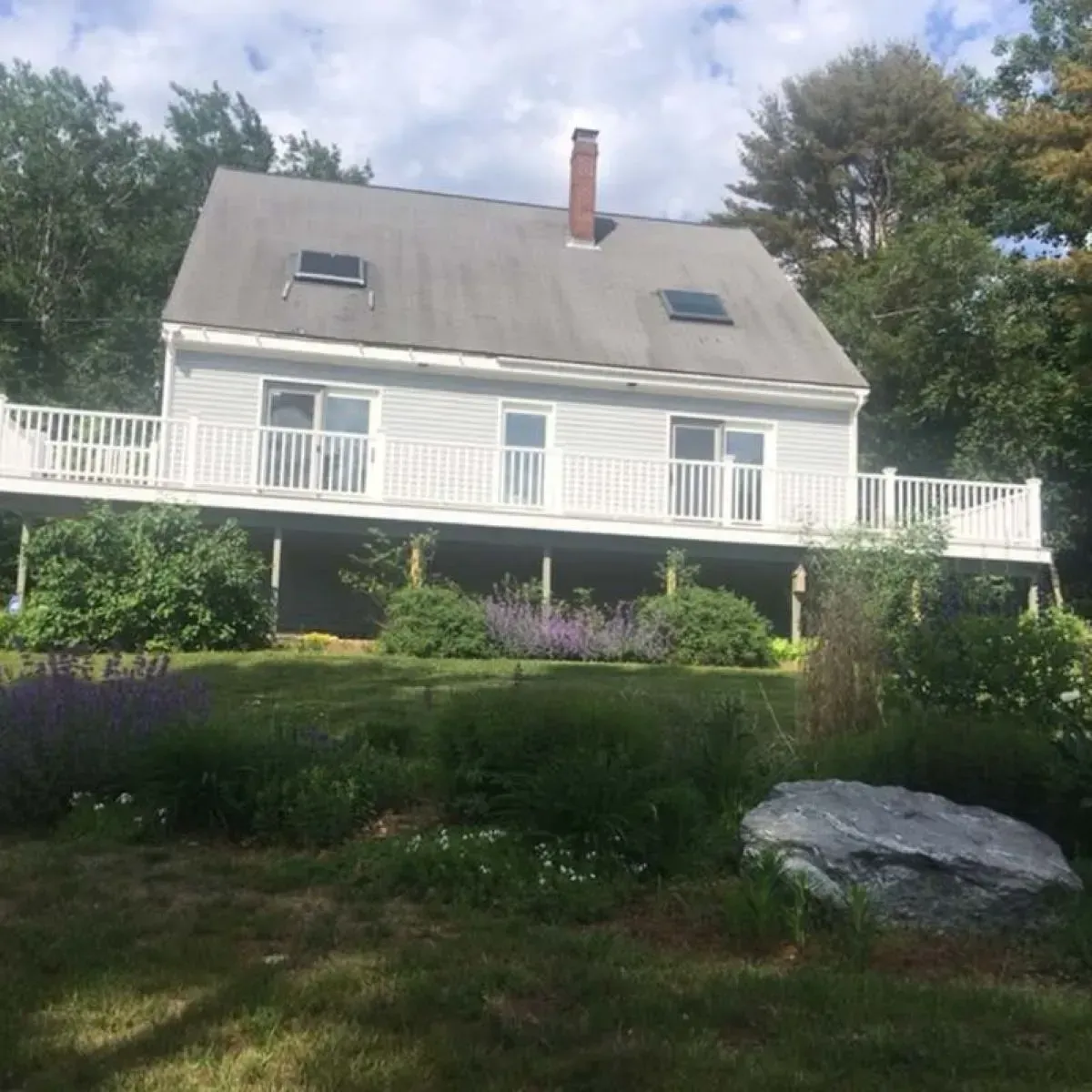 A white house with a large deck and a chimney surrounded by trees.
