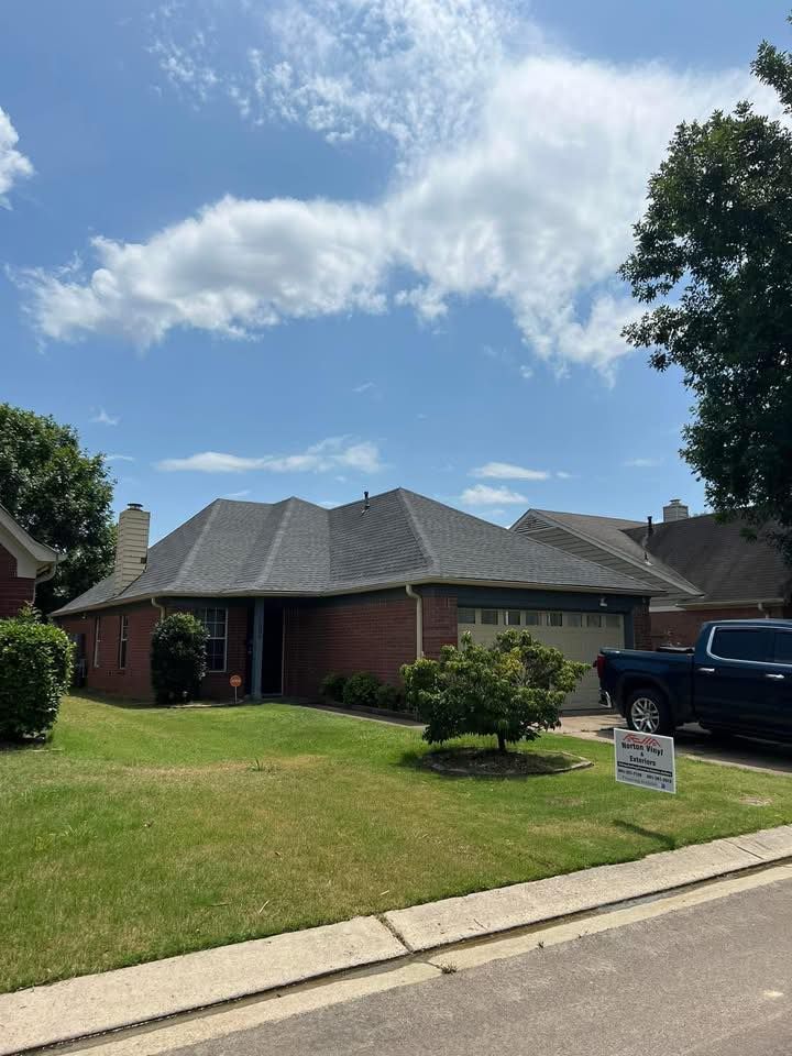Suburban brick house with gray roof, green lawn, and a pickup truck parked in the driveway under a partly cloudy sky
