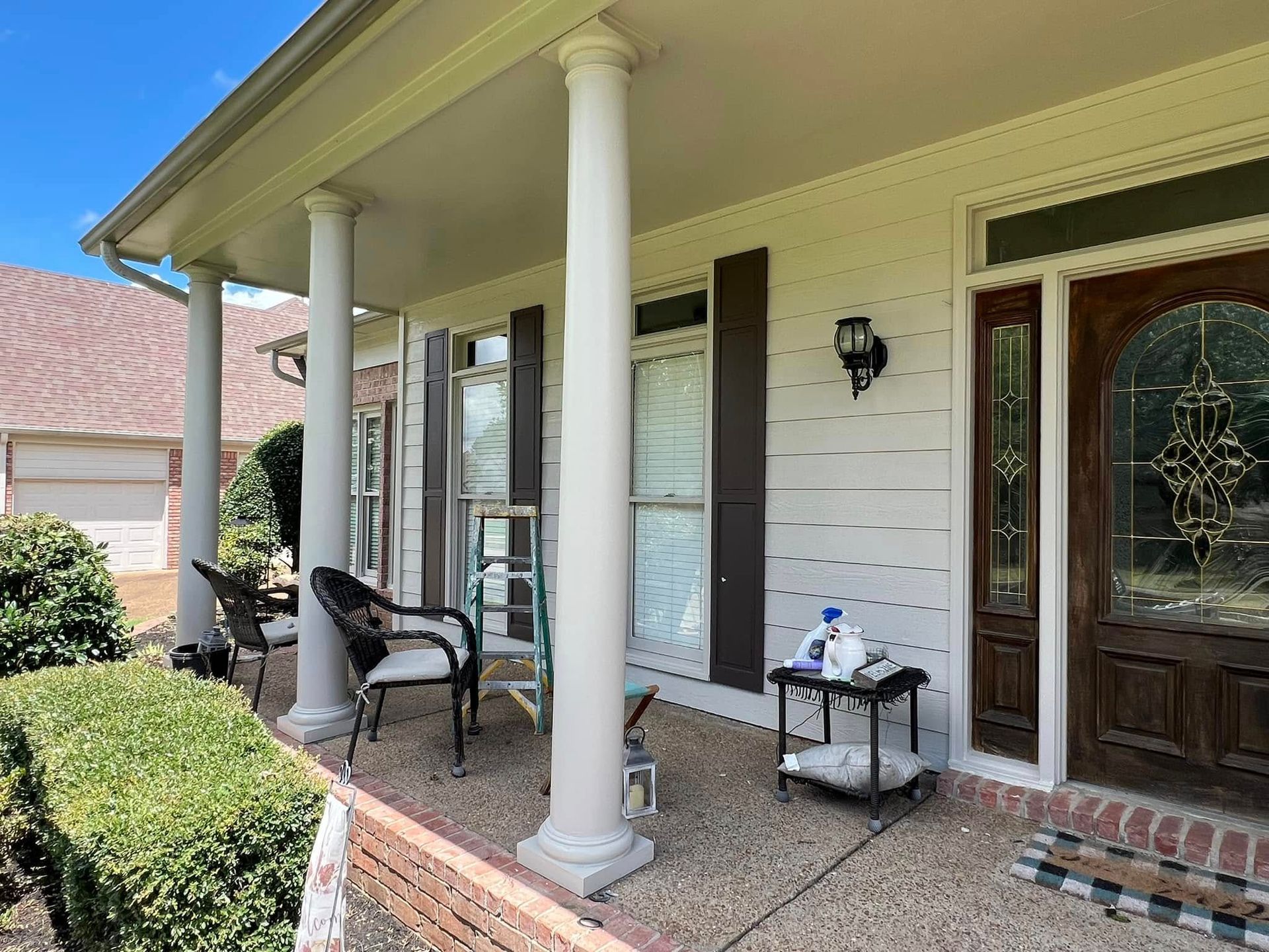 Front porch with white columns, wooden front door, chair, side table, and trimmed hedges by a brick house.