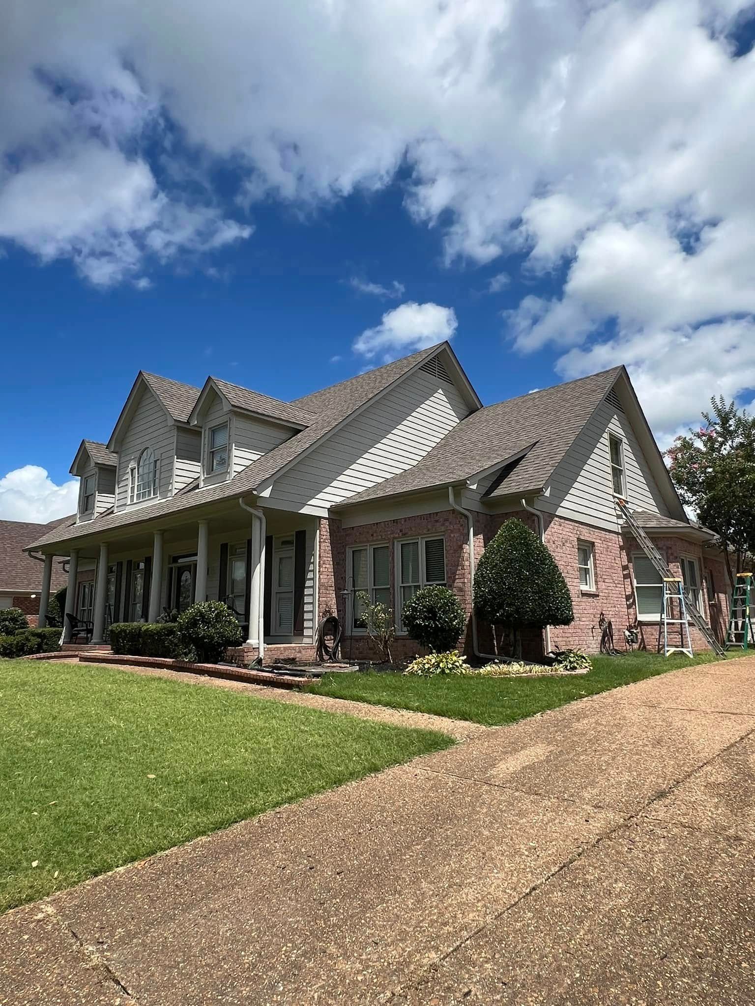 Large brick house with steep gabled roof, green lawn, and gravel driveway under a partly cloudy sky