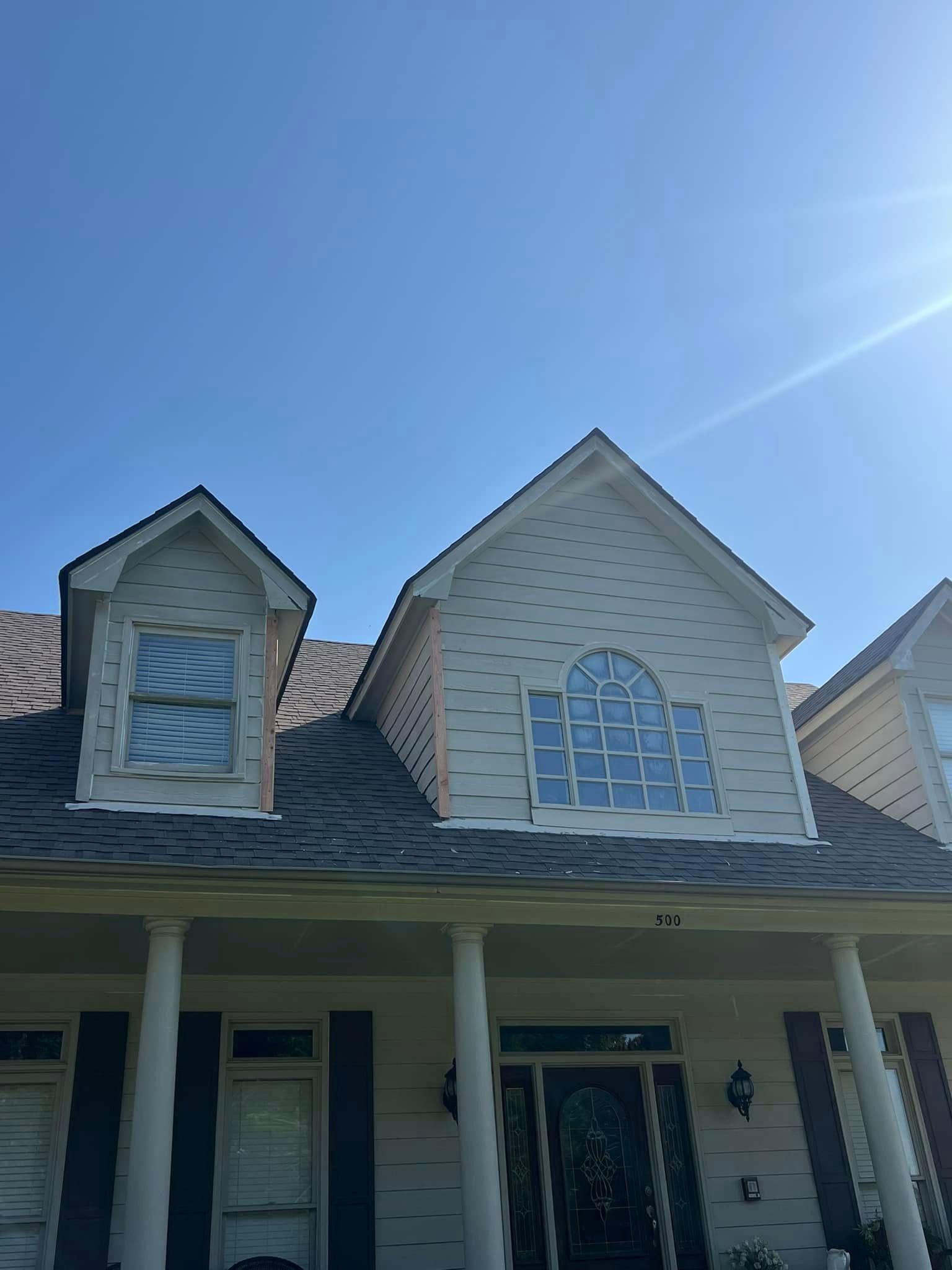 Suburban house exterior with beige siding, dormer windows, and a front porch under a bright blue sky
