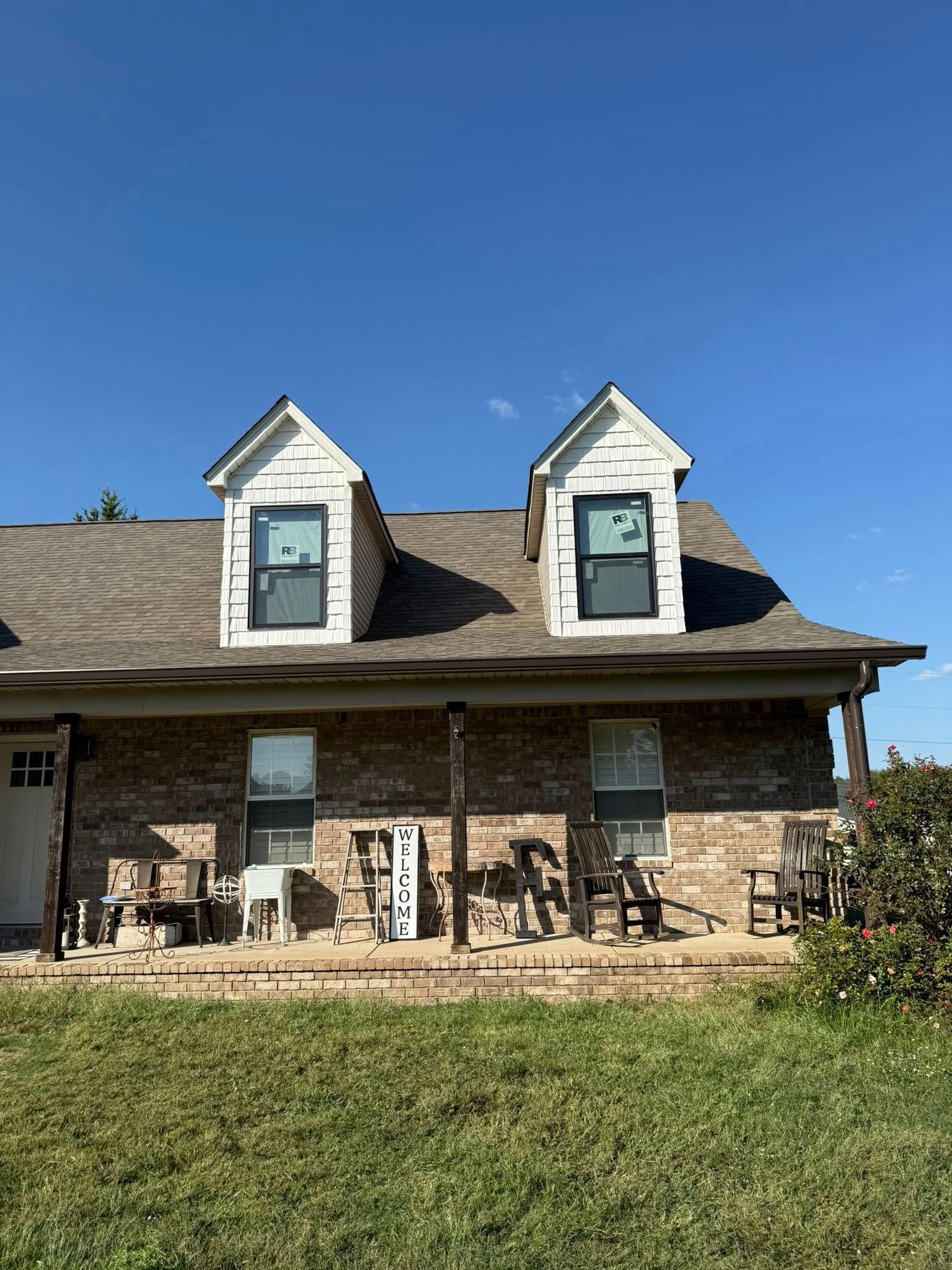 Brick house with two dormer windows, stone porch, and outdoor chairs under a blue sky