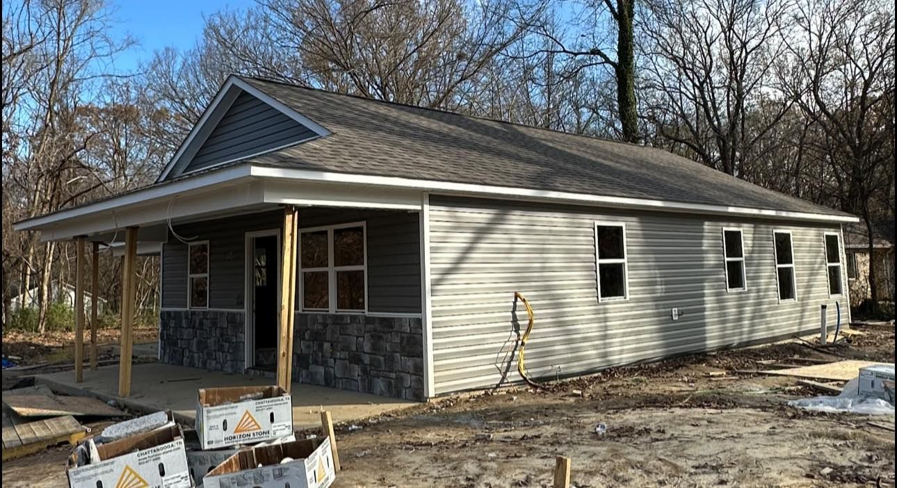 Single-story house under construction with beige siding, dark roof, and porch in a wooded yard
