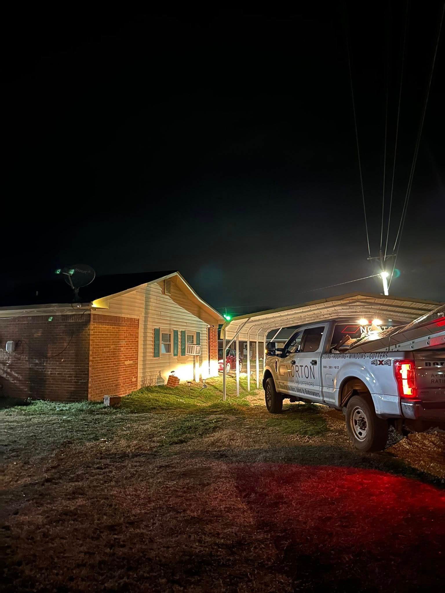 Nighttime rural scene with a lit brick house and a white pickup truck parked in front.