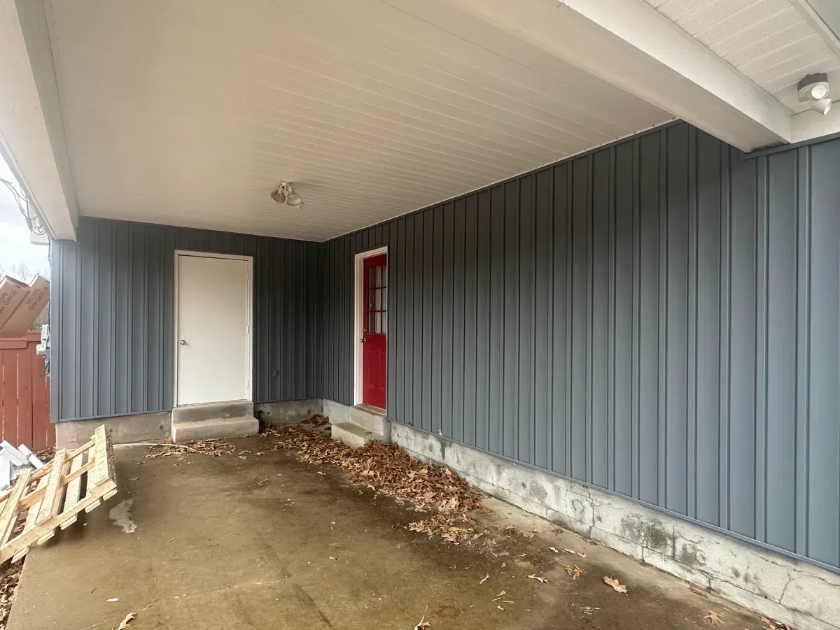Covered side porch with gray siding, red door, white utility door, and dirt floor under a slanted roof