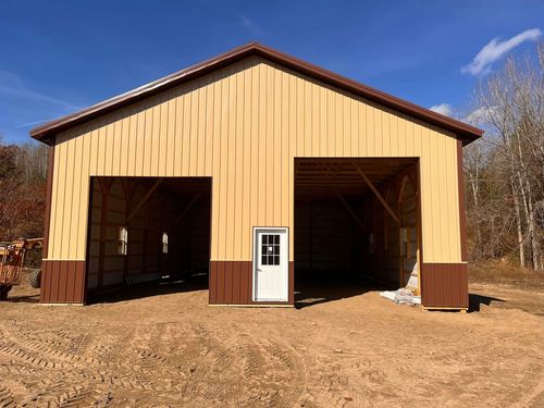 A new pole barn building with beige and brown metal siding and a white entry door, standing on a dirt lot.
