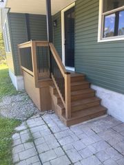 A set of five brown wooden steps with black metal railings leading to a dark door on a house with olive-green siding.